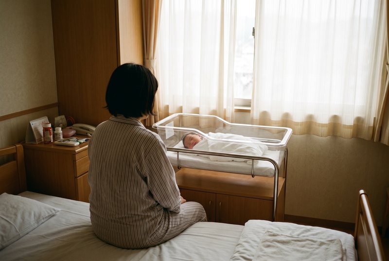 病室の窓辺で、赤ちゃんのベッドを見つめる母親